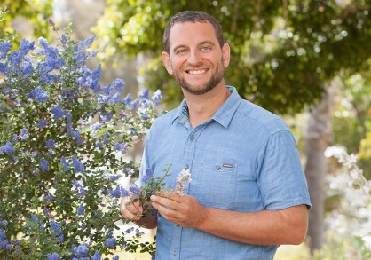 man in blue shirt holding a white flower next to a bush of blue flowers